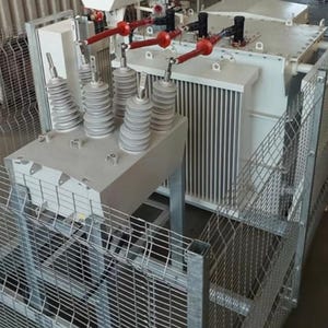 Puede incluir: Un gran transformador eléctrico industrial con una jaula protectora de metal. El transformador es principalmente blanco con componentes grises, incluyendo aisladores y conectores con puntas rojas. La jaula es de malla de alambre plateada.
