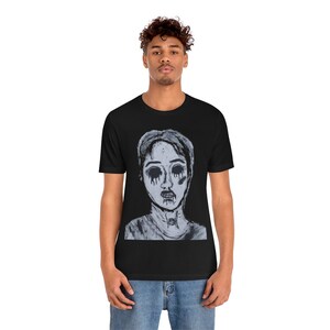 Puede incluir: Camiseta negra con una ilustración en escala de grises de una persona con cuencas oscuras y lágrimas negras goteando. La obra de arte tiene una estética gótica. La camiseta es de cuello redondo clásico.
