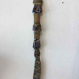 May include: A long, beaded Native American artifact with a tan suede-like body. The artifact is adorned with colorful beads and metal accents. The top has a leather loop, and the bottom has a beaded fringe.