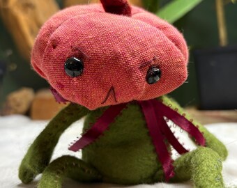 May include: A handmade plush toy frog with a red pumpkin-shaped head, black bead eyes, and a burgundy ribbon bow. The frog's body and limbs are green, and it is sitting on a white surface.