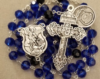 Puede incluir: Un rosario con cuentas facetadas azules y negras. El rosario presenta un crucifijo plateado, un escudo con la imagen de San Miguel y una placa de oficial de policía. El rosario está ensartado en una cadena plateada.