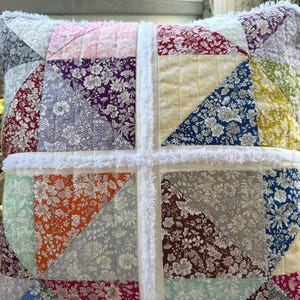 May include: Square patchwork pillow with a variety of floral patterned fabrics in shades of pink, purple, orange, blue, and gray. The pillow is trimmed with white fluffy fabric and has a white cross design.