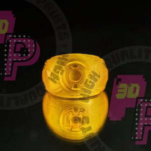 Pu&ograve; includere: Un anello giallo traslucido con un emblema circolare sulla parte anteriore. Il design include un motivo circolare centrale circondato da un bordo rialzato, creando un distinto motivo in rilievo. Il materiale appare liscio e lucido.