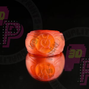 Pu&ograve; includere: Un anello arancione traslucido caratterizzato da un emblema circolare sulla parte anteriore con un simbolo centrale a forma di V circondato da un bordo circolare. L'anello ha una fascia liscia e arrotondata ed &egrave; mostrato con un riflesso.