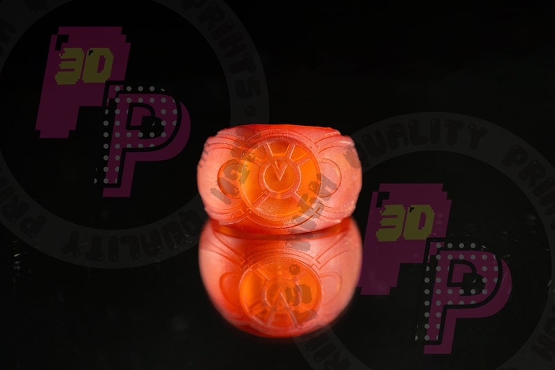 Pu&ograve; includere: Un anello arancione traslucido caratterizzato da un emblema circolare sulla parte anteriore con un simbolo centrale a forma di V circondato da un bordo circolare. L'anello ha una fascia liscia e arrotondata ed &egrave; mostrato con un riflesso.