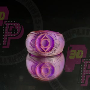 Pu&ograve; includere: Un anello viola con un emblema circolare caratterizzato da un cerchio centrale e due triangoli rivolti verso l'alto e verso il basso. L'anello presenta una finitura superficiale testurizzata e stratificata su uno sfondo scuro.