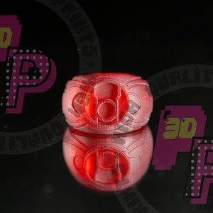 Pu&ograve; includere: Un anello rosso traslucido caratterizzato da un emblema circolare centrale con un design incassato. L'anello presenta una superficie strutturata e stratificata tipica della stampa 3D, con un simbolo prominente e un riflesso visibile sotto.