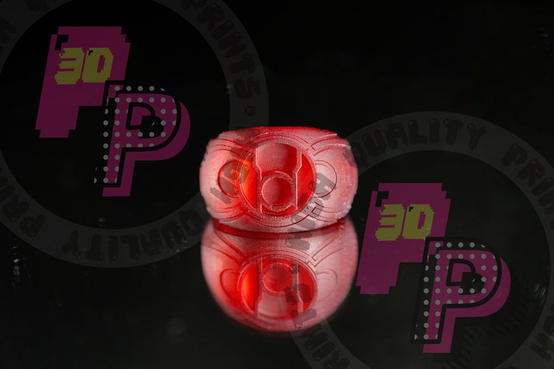 Pu&ograve; includere: Un anello rosso traslucido caratterizzato da un emblema circolare centrale con un design incassato. L'anello presenta una superficie strutturata e stratificata tipica della stampa 3D, con un simbolo prominente e un riflesso visibile sotto.