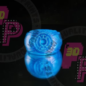 Pu&ograve; includere: Un anello blu brillante caratterizzato da un emblema circolare con un punto centrale e quattro linee radianti sulla parte frontale. L'anello presenta una fascia liscia e arrotondata con una finitura lucida che riflette la luce su sfondo scuro.
