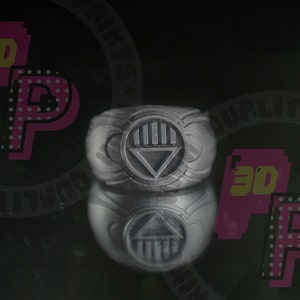 Pu&ograve; includere: Un anello grigio opaco con un emblema circolare in rilievo sulla parte anteriore. L'emblema mostra un design a barre verticali sopra una forma triangolare, caratteristico del simbolo del Black Lantern Corps. L'anello ha una finitura metallica testurizzata.