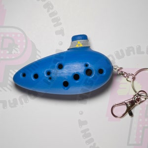 May include: A blue ocarina keychain with a silver key ring. The ocarina has a triangular design on the top and is made of plastic.