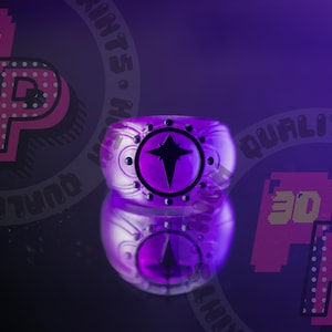 Può includere: Un anello viola caratterizzato da una faccia circolare con un emblema a stella nera a quattro punte al centro. La fascia presenta un design testurizzato con piccoli accenti circolari neri che circondano il motivo centrale a stella, in una tonalità viola.