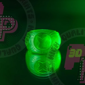 Op de afbeelding: Een groene plastic ring met een gloeiend groen symbool in het midden. De ring ligt op een groene ondergrond met een roze en geel logo op de achtergrond.