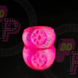 Pu&ograve; includere: Un anello rosa vibrante e traslucido con un prominente emblema a forma di stella sulla parte anteriore. La fascia presenta un design in rilievo e testurizzato sui lati. L'anello &egrave; posizionato in modo da mostrare chiaramente il profilo frontale.