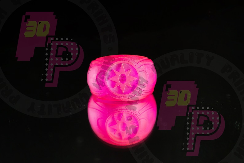 Pu&ograve; includere: Un anello rosa vibrante e traslucido con un prominente emblema a forma di stella sulla parte anteriore. La fascia presenta un design in rilievo e testurizzato sui lati. L'anello &egrave; posizionato in modo da mostrare chiaramente il profilo frontale.