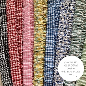 May include: A collection of fabric badge reels in various patterns and colors, including gingham, floral, and solid colors. The reels are arranged vertically, showcasing a variety of designs. Text on the image indicates "+20 prints", "breakaway option", and "MRI safe badge reel".