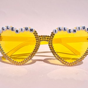 Puede incluir: Gafas de sol amarillas en forma de corazón con pedrería y la palabra "ILLENIUM" escrita en cuentas blancas.