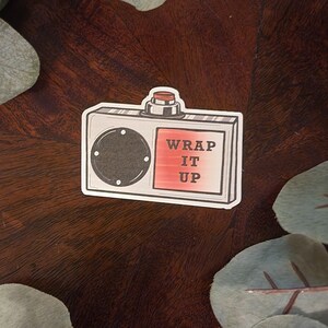 May include: A white and red sticker with a cartoon illustration of a vintage radio with a red button. The radio has a black dial and the text "WRAP IT UP" on the front.