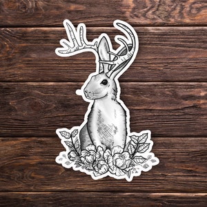 May include: A black and white illustration of a rabbit with antlers and flowers. The rabbit is sitting on a bed of flowers and has a whimsical, fairytale-like quality.