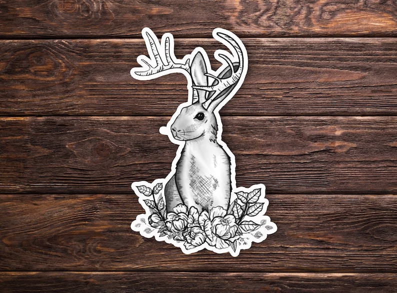 May include: A black and white illustration of a rabbit with antlers and flowers. The rabbit is sitting on a bed of flowers and has a whimsical, fairytale-like quality.