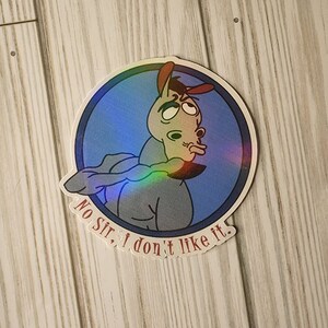 May include: A round sticker with a holographic finish features a cartoon donkey with a red hat. The donkey is looking to the side with a disapproving expression. The text "No Sir, I don't like it." is written in a red font around the bottom of the sticker.