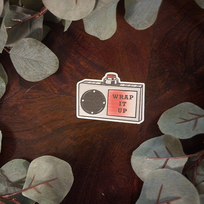 May include: A white and red sticker with a cartoon illustration of a vintage radio with a red button. The radio has a black dial and the text "WRAP IT UP" on the front.