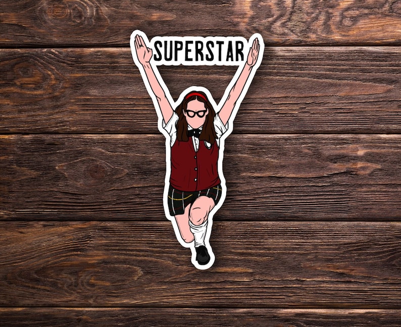 Puede incluir: Una pegatina con una ilustraci&oacute;n de dibujos animados de una persona con chaleco rojo, falda a cuadros y gafas, con los brazos levantados. La palabra "SUPERSTAR" est&aacute; encima de la cabeza de la figura. Fondo de madera oscura.