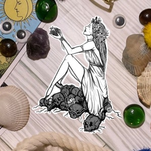 May include: A black and white illustration of a woman with a flower crown sitting on a pile of skulls. She is holding a heart in her hand and looking up. The image is surrounded by a white border.