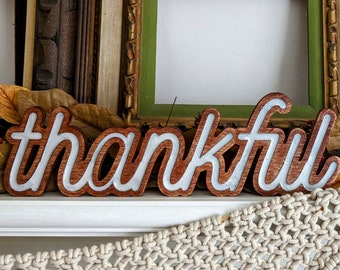 May include: A wooden sign with the word "thankful" in cursive script, with a white outline. The sign is placed in front of vintage picture frames and fall foliage. The frames have gold and green accents.