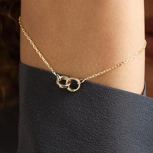 May include: A delicate gold bracelet featuring two interlocking circles. The bracelet is worn on a wrist, with a black long-sleeved shirt in the background. The jewelry is simple and elegant.