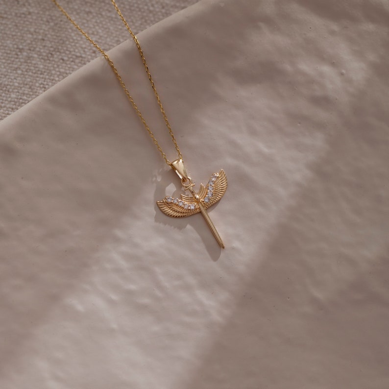 May include: A gold necklace with a pendant featuring a sword with wings and clear crystals.