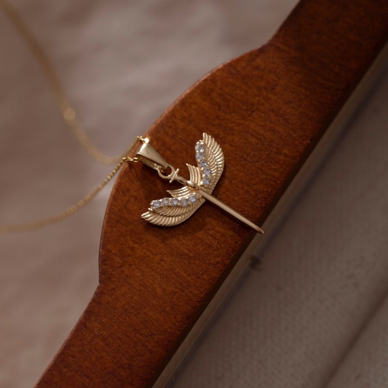 May include: A gold pendant necklace with a sword design featuring clear crystal accents. The sword is surrounded by two wings, also adorned with crystals.
