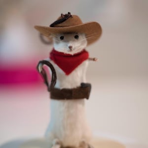 May include: A white mouse wearing a brown cowboy hat, a red bandana, and a brown belt. The mouse is standing on a white surface.