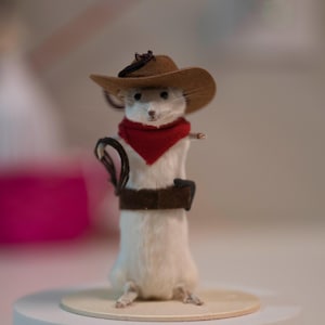 May include: A small white mouse wearing a brown cowboy hat, a red bandana, and a brown belt. The mouse is standing on a wooden platform.