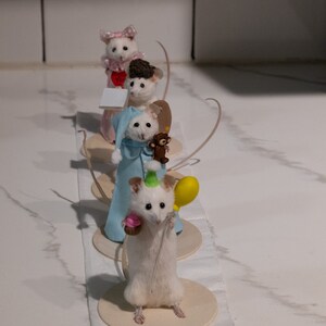 May include: Four white mouse figurines wearing different outfits and accessories. The mouse in the front is wearing a party hat and holding a cupcake and a yellow balloon. The mouse behind it is wearing a blue robe and holding a teddy bear. The mouse behind that is wearing a brown hat and the mouse in the back is wearing a pink dress and a bow.