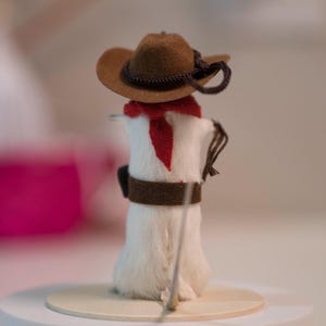 May include: A white, furry cowboy figurine wearing a brown felt hat with a black band, a red bandana, and a brown leather belt. The figurine is holding a stick in its right hand.