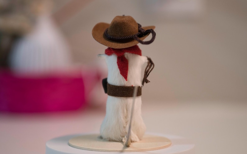 May include: A white, furry cowboy figurine wearing a brown felt hat with a black band, a red bandana, and a brown leather belt. The figurine is holding a stick in its right hand.