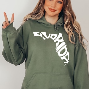 May include: A green hooded sweatshirt with a white outline of the state of Florida, with the word "Florida" spelled out within the outline. The model is wearing the sweatshirt and is making a peace sign with her left hand.