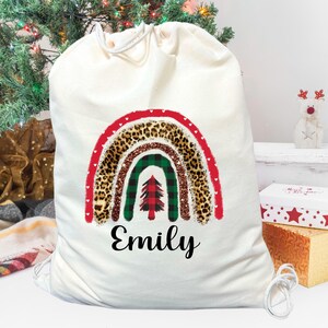 May include: A natural canvas Christmas gift bag with a rainbow design in red, green, and leopard print. The bag has the name "Emily" in black script. A Christmas tree and presents are in the background.