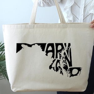 May include: A cream-colored tote bag with black graphic text and a map outline of Maryland. The bag has long, sturdy straps for carrying and is made of a durable fabric.