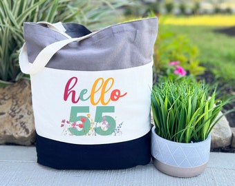 May include: A grey and white tote bag with the text "hello 55" in colorful letters and a floral design. The bag has a black bottom and a white top with a grey stripe.