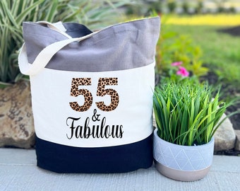 May include: A tri-color tote bag with a white panel featuring the number 55 in a leopard print design and the text "& Fabulous". The bag is grey, white, and black.