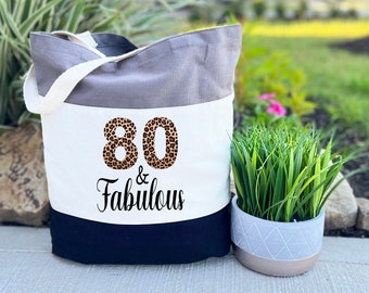 May include: A tri-color tote bag with a white panel featuring the text "80 & Fabulous" in black with the number 80 in a leopard print design. The bag is grey, white, and black.
