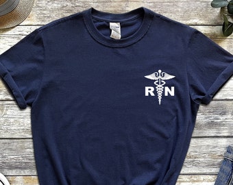 以下が含まれることがあります： 白い医療シンボルと「RN」の文字が書かれたネイビーブルーのTシャツ。