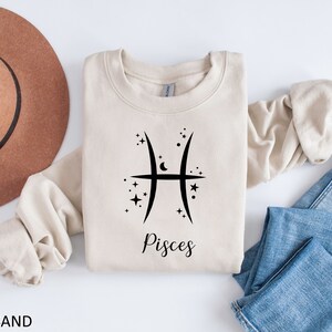Op de afbeelding: Een beige sweatshirt met een zwart Vissen-sterrenbeeld en het woord "Vissen" bedrukt op de voorkant. Het sweatshirt is gevouwen en wordt getoond op een wit oppervlak met een bruine hoed en een blauwe spijkerbroek.