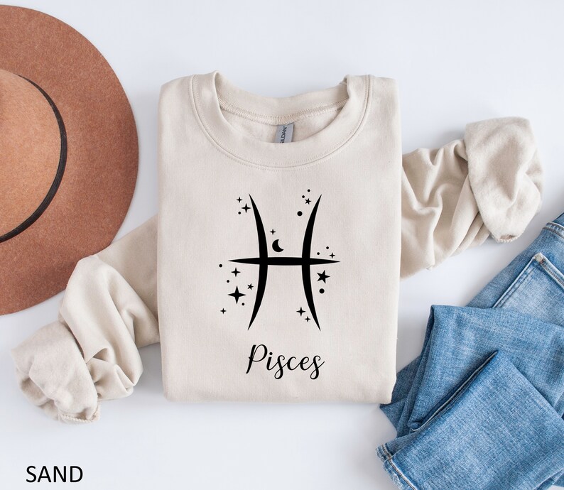 Op de afbeelding: Een beige sweatshirt met een zwart Vissen-sterrenbeeld en het woord "Vissen" bedrukt op de voorkant. Het sweatshirt is gevouwen en wordt getoond op een wit oppervlak met een bruine hoed en een blauwe spijkerbroek.