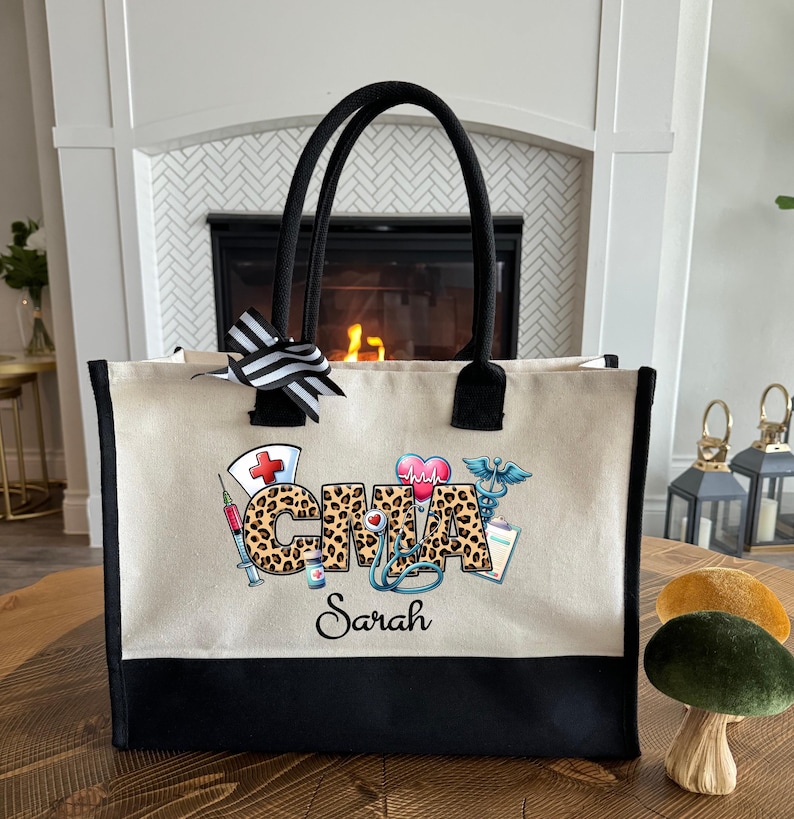 K&ouml;nnte beinhalten: Beige Tragetasche mit schwarzen Verzierungen und Griffen. Die Tasche zeigt die Buchstaben "CMA" in einem Leopardenmuster, zusammen mit medizinischen Grafiken und dem Namen "Sarah". Eine schwarz-wei&szlig; gestreifte Schleife ziert die Oberseite.