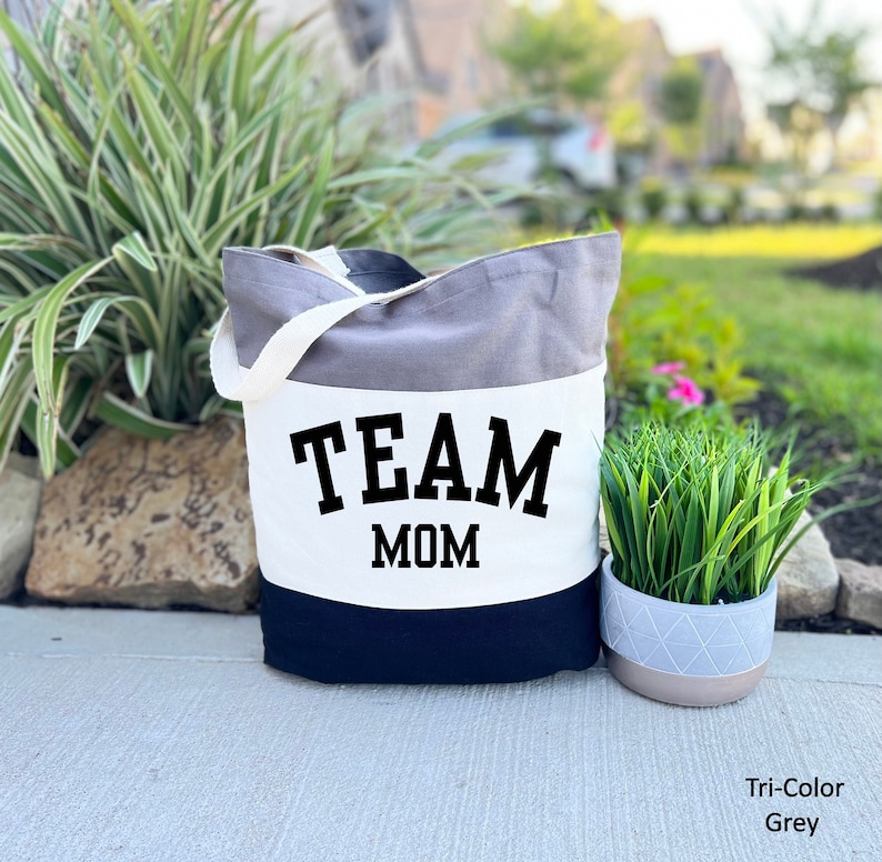 May include: A gray, white, and black canvas tote bag with the text "TEAM MOM" in black. The bag has a white strap.