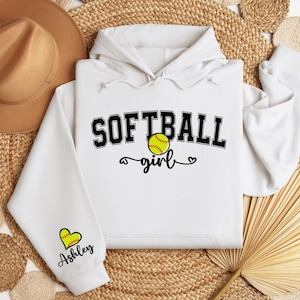 May include: White hoodie with a black graphic that says "Softball girl" with a softball graphic and a heart. The sleeve has a heart graphic with the name "Ashley" written below it.