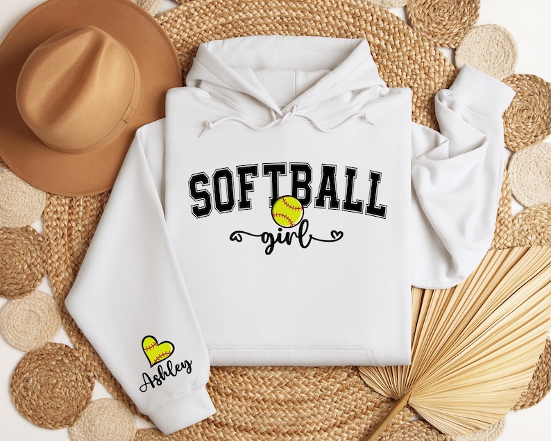 May include: White hoodie with a black graphic that says "Softball girl" with a softball graphic and a heart. The sleeve has a heart graphic with the name "Ashley" written below it.